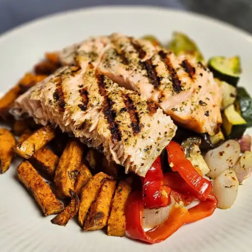 Gegrillter Lachs mit Süßkartoffelpommes und Ofengemüse auf einem Teller angerichtet