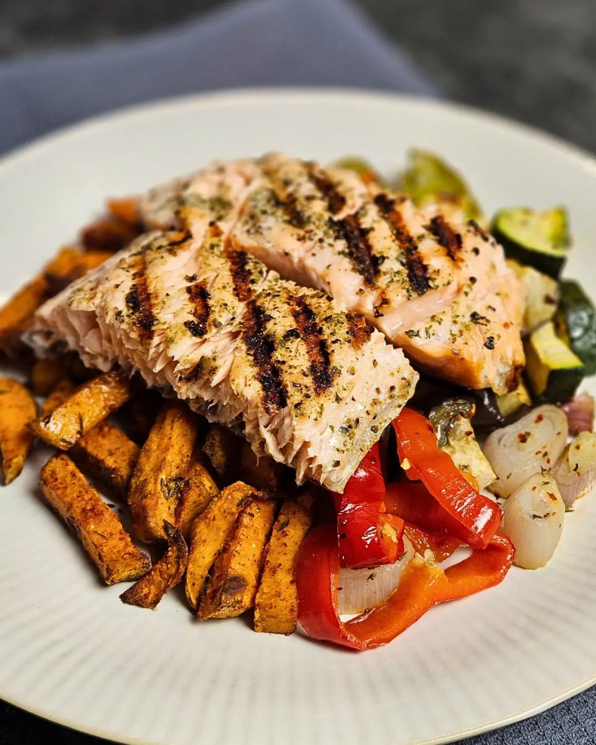 Gegrillter Lachs mit Süßkartoffelpommes und Ofengemüse auf einem Teller angerichtet