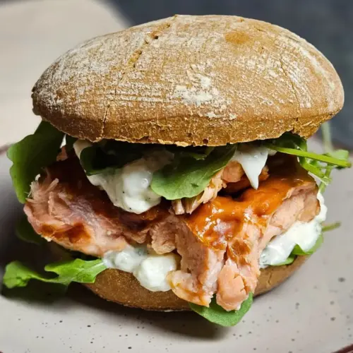 Pulled Salmon Burger mit Rucola und cremiger Gurken-Quark-Sauce auf einem Teller angerichtet