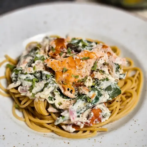 Pulled Salmon Pasta mit cremiger Frischkäse-Spinat-Sauce auf einem Teller mit einem Salat im Hintergrund