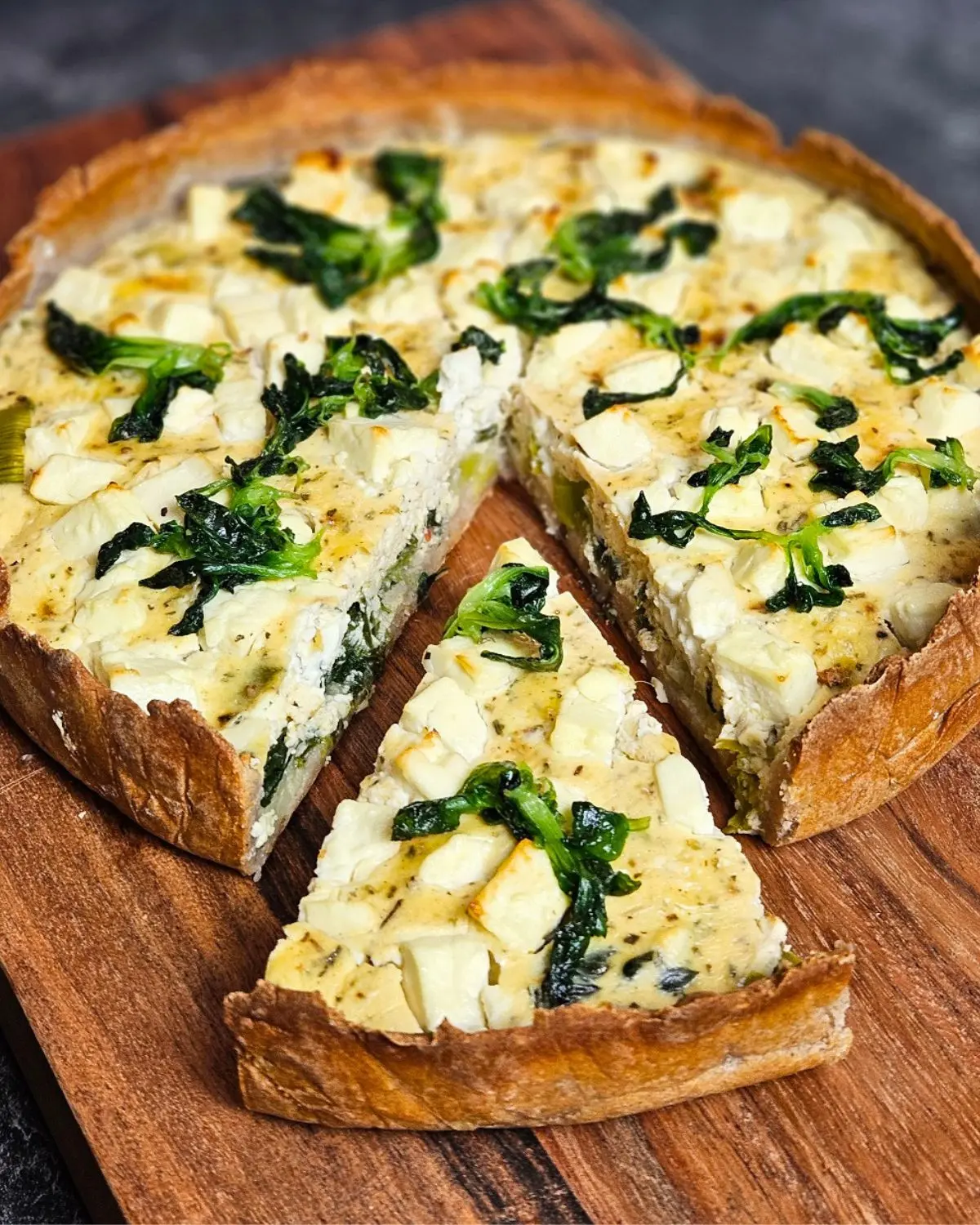 Spinat-Feta-Quiche mit Lauch aus Quarkteig auf einem Brett angerichtet, ein Stück ist rausgeschnitten