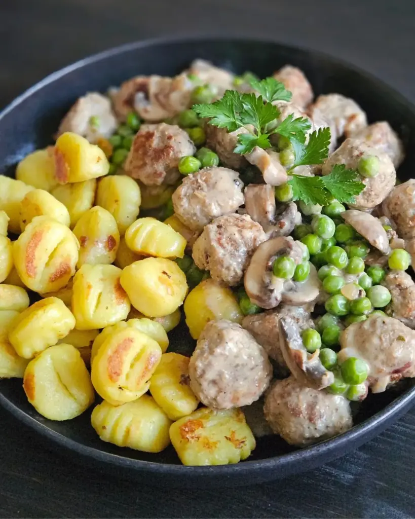 Gnocchi mit Hackbällchen, Champignons und Erbsen auf einem schwarzen Teller angerichtet