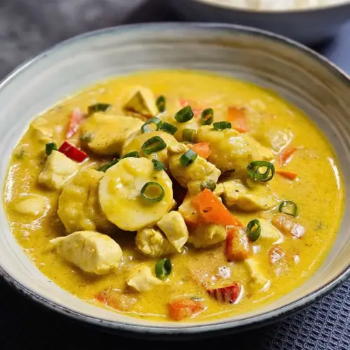 Karibisches Hähnchencurry mit Kokosmilch und Banane in einer Schüssel angerichtet mit einer Schüssel Reis im Hintergrund