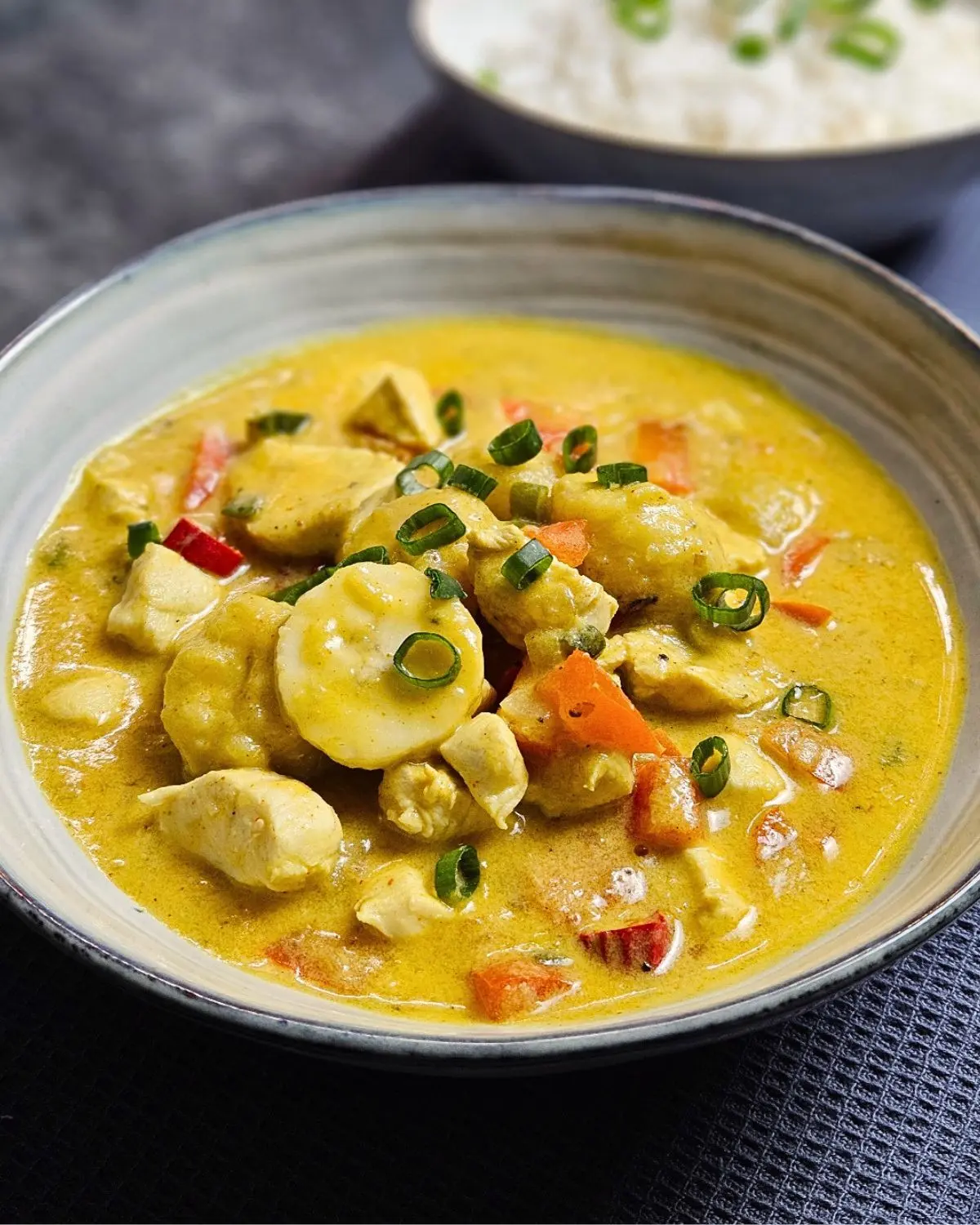 Karibisches Hähnchencurry mit Kokosmilch und Banane in einer Schüssel angerichtet mit einer Schüssel Reis im Hintergrund